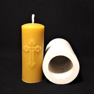 May include: A yellow beeswax candle with a cross design and a white wick. The candle is next to a white silicone mold for making candles.