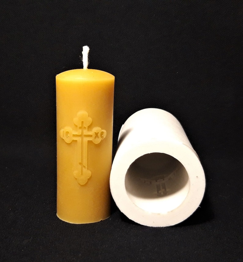 May include: A yellow beeswax candle with a cross design and a white wick. The candle is next to a white silicone mold for making candles.