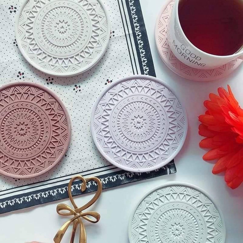 May include: Four round coasters with a mandala design. The coasters are in shades of grey, pink, and white. The coasters are made of silicone and have a raised design. The coasters are on a white and black patterned surface. The text "Good Morning" is on a pink coaster.