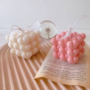 Peut inclure: Deux bougies carrées avec un motif en forme de cœur. Une bougie est blanche et l'autre est rose.