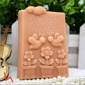 May include: A light brown soap bar with a bee theme. The soap has the text "Let's bee friends..." and features two bees, flowers, and pots.