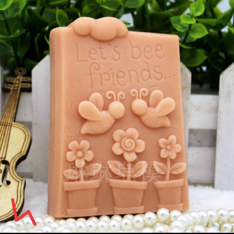 May include: A light brown soap bar with a bee theme. The soap has the text "Let's bee friends..." and features two bees, flowers, and pots.