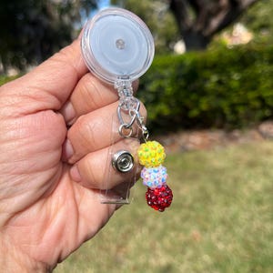 May include: A clear retractable badge reel with a decorative charm. The charm features three stacked, sparkling beads in yellow, white, and red. The reel has a clear plastic strap and a metal clip.