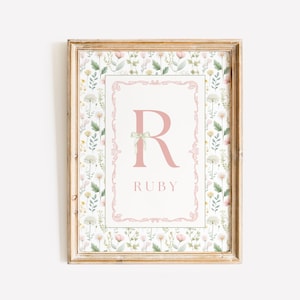 May include: A framed print featuring a floral pattern background with a pink border. The print displays the letter "R" and the name "RUBY" in pink. The frame is a light brown color.
