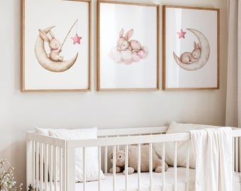 May include: Three framed watercolour prints. Each print features a sleeping bunny on a crescent moon, a bunny on a cloud, and a bunny on a crescent moon with a star. The prints are in light brown wooden frames above a white cot.