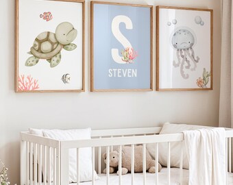 May include: Three framed watercolor art prints with a nautical theme. One features a turtle, another a jellyfish, and the third a blue background with the letter "S" and the name "STEVEN". The prints are in wooden frames and hang above a white crib.