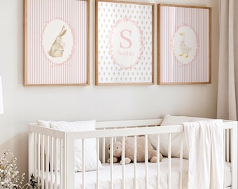 May include: Three framed nursery art prints. One features a bunny, another a monogram with the name "Sophia", and the third a goose. All are in oval frames with pink accents and set against a striped or patterned background. The frames are wood.
