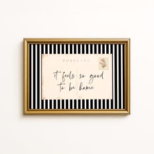 May include: A framed print with a gold border. The print features a black and white striped background and a vintage postcard with the text "it feels so good to be home." A floral stamp is in the upper right corner.