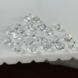 Peut inclure: Une collection de cristaux de quartz clairs et facetés, également appelés diamants de Herkimer, sur une surface blanche.