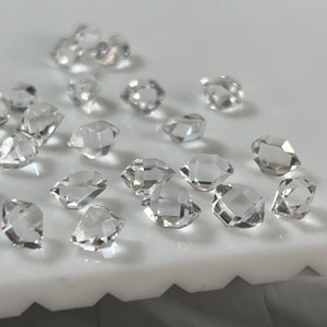 May include: A collection of clear, faceted, quartz crystals, also known as Herkimer diamonds, on a white surface.