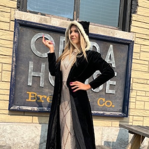 May include: A woman wearing a black velvet coat over a sequined dress. The dress has a geometric pattern in shades of gold and beige. She is also wearing a black hat with a white fur trim. The sign behind her says "Opera House Brewing Co."
