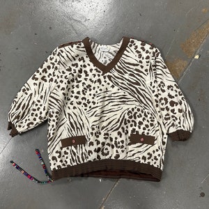 Puede incluir: Una blusa de estampado animal marrón y blanco con cuello en V y mangas 3/4. La blusa tiene dos bolsillos en la parte delantera y una cintura elástica marrón.