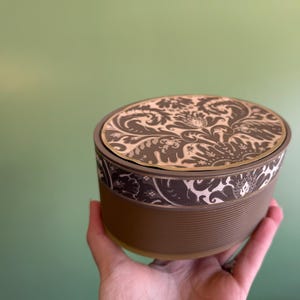 May include: A round, brown container with a patterned lid. The lid has a gold trim and a black and white floral design. The container has a ribbed texture.