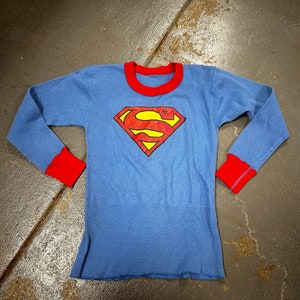 May include: Blue long-sleeved thermal shirt with red trim on the collar and cuffs. The shirt features the Superman logo in the center, a red diamond with a yellow 'S' inside. The shirt is made of a waffle-knit fabric.