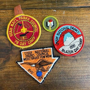May include: A collection of vintage Utah National Parks Council patches. The patches feature various designs, including a red and yellow circular patch with text and a logo, and a triangular patch with an eagle.