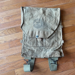 Könnte beinhalten: Ein beigefarbener Rucksack aus Segeltuch mit einer großen oberen Klappe, einem kreisförmigen Emblem und einem einzelnen Metalldruckknopf. Die Tasche verfügt über zwei Vordertaschen und zwei verstellbare olivgrüne Gurtbänder mit schwarzen Metallclips.