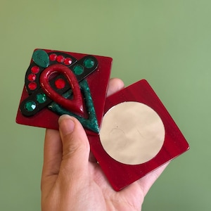 May include: A red square compact with a decorative top featuring red, green, and black accents. The open compact reveals a round mirror. The design includes red and green jewels.
