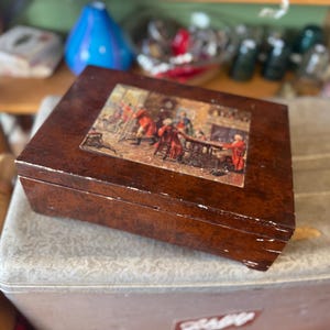 Op de afbeelding: Een vintage houten kist met een scharnierend deksel. De kist is donkerbruin van kleur met een decoratieve afbeelding op de bovenkant. De afbeelding toont een scène met mensen in historische kleding. De kist staat op een grijze ondergrond.