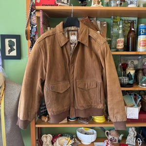 Può includere: Un giubbotto bomber in pelle color cuoio con colletto classico, maniche lunghe con polsini a coste, orlo a coste e due ampie tasche frontali con patta. La giacca è appesa a una gruccia nera e presenta un'etichetta interna visibile al collo.