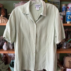 Puede incluir: Una camisa verde claro con botones y mangas cortas, colgada de una percha. La camisa tiene cuello y botones en la parte delantera. La marca "PENDLETON" es visible en una etiqueta cerca del cuello.
