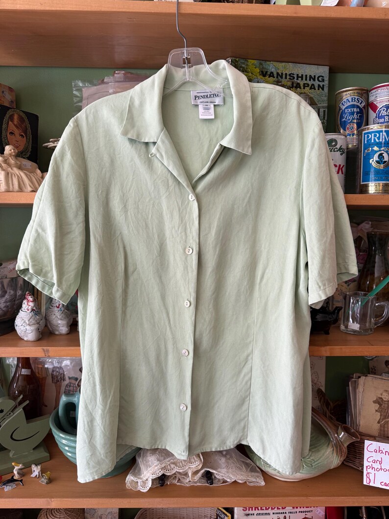 Puede incluir: Una camisa verde claro con botones y mangas cortas, colgada de una percha. La camisa tiene cuello y botones en la parte delantera. La marca "PENDLETON" es visible en una etiqueta cerca del cuello.