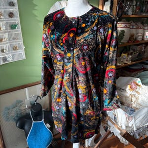 Puede incluir: Vestido de manga larga con botones y un vibrante estampado abstracto en rojo, amarillo, azul y naranja sobre fondo negro. El vestido se exhibe en un maniquí blanco.