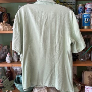 Puede incluir: Una camisa verde claro de manga corta con cuello, colgada de una percha met&aacute;lica. La camisa tiene una costura vertical en la espalda y est&aacute; hecha de una tela ligera. El fondo presenta estantes con varios art&iacute;culos vintage y el texto "VANISHING JAPAN".