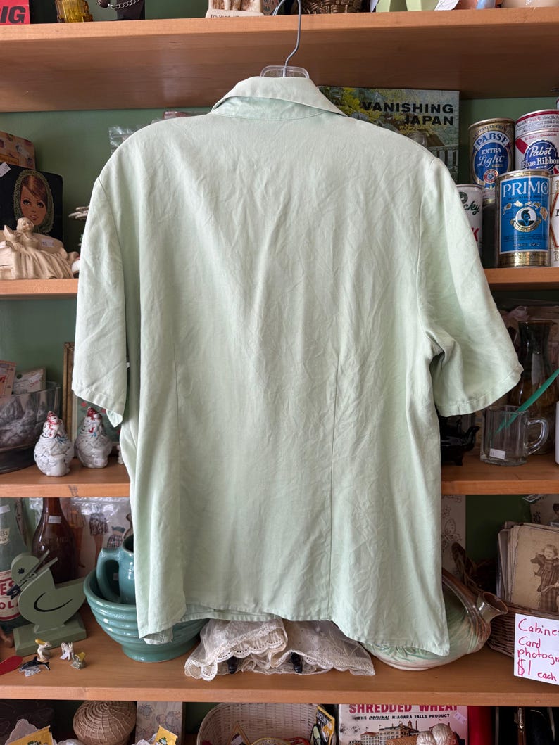 Puede incluir: Una camisa verde claro de manga corta con cuello, colgada de una percha met&aacute;lica. La camisa tiene una costura vertical en la espalda y est&aacute; hecha de una tela ligera. El fondo presenta estantes con varios art&iacute;culos vintage y el texto "VANISHING JAPAN".