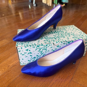 May include: A pair of cobalt blue satin pumps with pointed toes and low heels. The shoes are displayed on a patterned box with the brand name visible. The shoes are on a wooden surface.