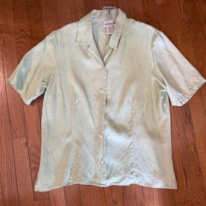 Puede incluir: Una camisa verde claro de manga corta con botones. La camisa tiene cuello y una textura sutil. Est&aacute; hecha de una tela ligera y tiene un corte relajado. La camisa est&aacute; colgada en una percha.