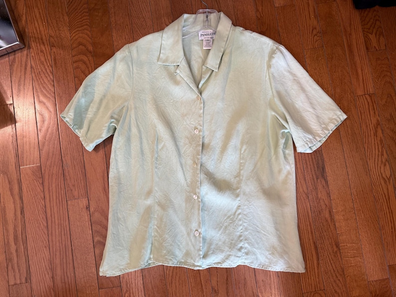 Puede incluir: Una camisa verde claro de manga corta con botones. La camisa tiene cuello y una textura sutil. Est&aacute; hecha de una tela ligera y tiene un corte relajado. La camisa est&aacute; colgada en una percha.