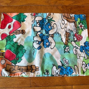 Pode incluir: Um tecido retangular com uma impressão colorida dos Smurfs numa cena florestal caprichosa. O design inclui personagens Smurf azuis, casas de cogumelos vermelhos e brancos e várias folhagens verdes sobre um fundo azul claro e creme.