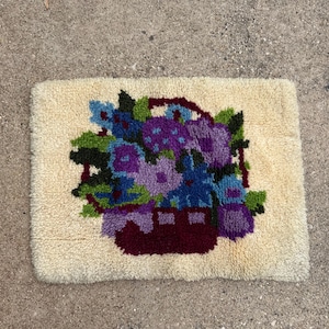 May include: A white rug with a colorful floral design. The rug features a basket filled with purple, blue, and green flowers. The basket is a dark red color.