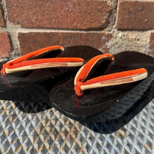 May include: A pair of black geta sandals with orange and beige straps. The sandals are traditional Japanese footwear.