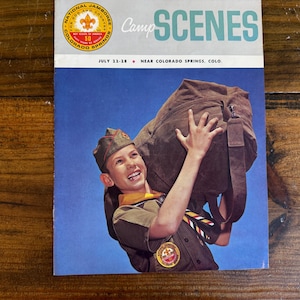 May include: Vintage magazine cover featuring a smiling boy in a Boy Scout uniform carrying a large brown duffel bag. The magazine title reads "Camp Scenes" with text indicating the National Jamboree in Colorado Springs.