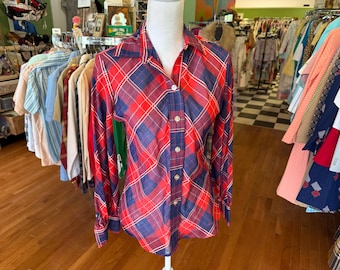May include: A red, white, and blue plaid button-down shirt with long sleeves. The shirt is displayed on a mannequin in a vintage clothing store. The shirt has a classic collar and buttoned cuffs.