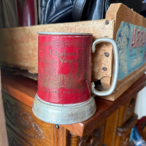 May include: A vintage red mug with a silver handle and base. The mug features gold lettering that reads "Las Vegas" and "Grand Central". The mug is sitting on a wooden surface.