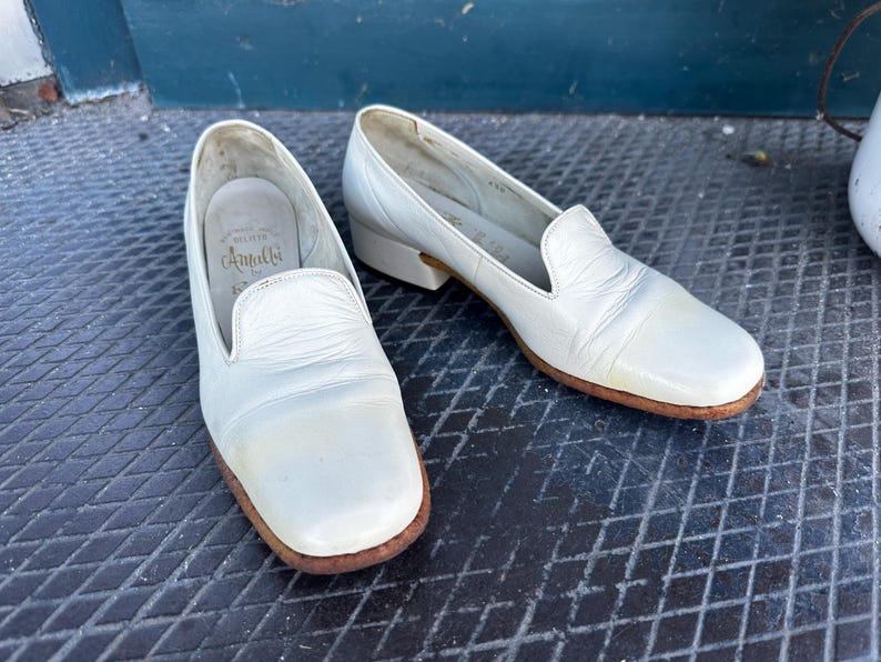 Pu&ograve; includere: Un paio di scarpe da donna in pelle bianca con punta arrotondata. Le scarpe hanno un design semplice e sono realizzate in pelle morbida e flessibile. Il nome del marchio "Amalfi" &egrave; visibile sulla soletta di una scarpa.