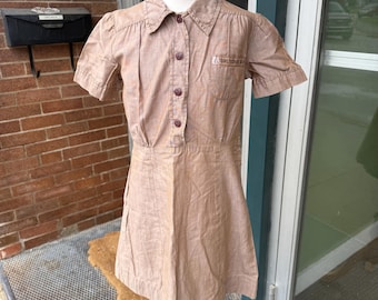 May include: A vintage brown Girl Scout uniform dress with short sleeves, a collared neckline, and a button-down front. The dress features a small pocket and the words "Girl Scouts" on the chest. The dress is displayed on a mannequin.