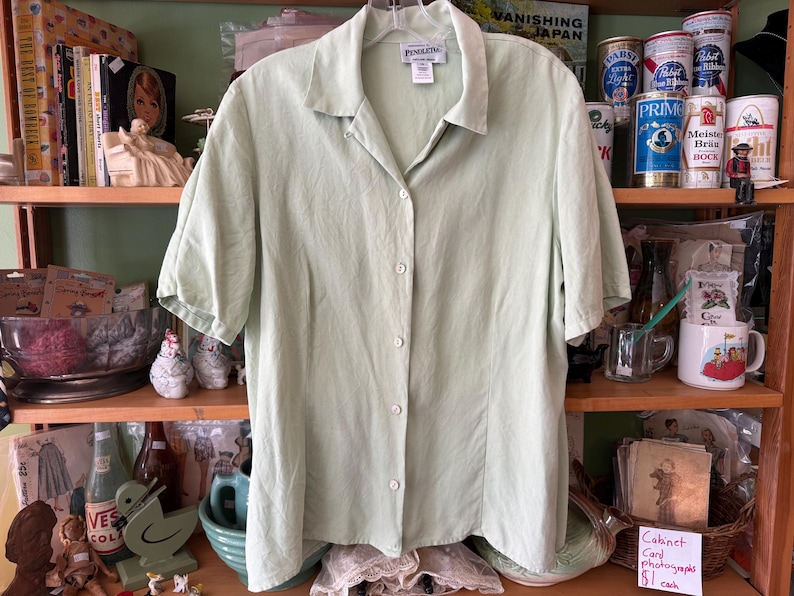 Puede incluir: Una camisa verde claro de manga corta con botones. La camisa tiene cuello y est&aacute; hecha de una tela ligera. Se exhibe en una estanter&iacute;a de madera con varios art&iacute;culos vintage.