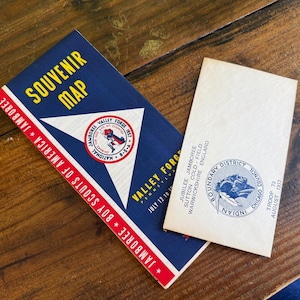 May include: A vintage souvenir map and a printed card. The map is blue with yellow text and a red and white border, featuring a Jamboree logo. The card has a circular emblem and text.