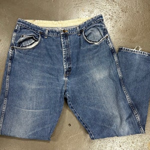 May include: Blue denim jeans with a classic five-pocket design. The jeans have a faded wash and a straight leg fit. The waistband is light-colored, and the jeans are laid flat on a concrete surface.