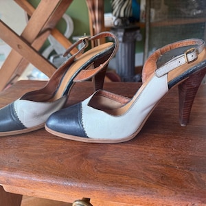 May include: A pair of two-tone slingback heels featuring a dark navy blue rounded toe cap with scalloped edges, a cream-coloured body, and a brown stacked high heel. The shoes include an adjustable ankle strap with a small gold-toned metal buckle.