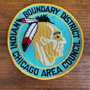 May include: A circular embroidered patch with a turquoise background and a yellow border. It features a profile illustration of a person wearing a headdress with white, red, and black feathers. The text reads Indian Boundary District Chicago Area Council.