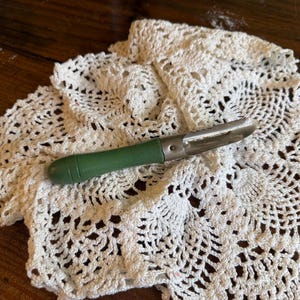 Puede incluir: Un pelador de verduras vintage con un mango verde y una cuchilla de acero inoxidable descansa sobre un tapete blanco de ganchillo. El tapete tiene un diseño intrincado y calado. El fondo es una superficie de madera oscura.