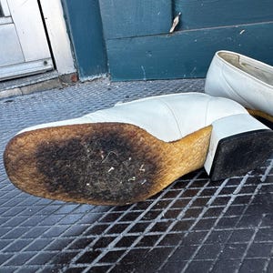 Pu&ograve; includere: Un paio di scarpe in pelle bianca con una suola spessa e usurata. Le scarpe hanno un tacco a blocco e una punta a punta.