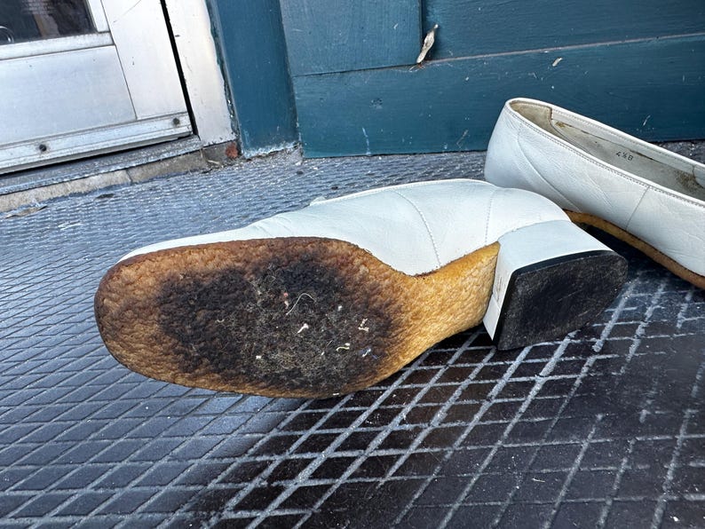 Pu&ograve; includere: Un paio di scarpe in pelle bianca con una suola spessa e usurata. Le scarpe hanno un tacco a blocco e una punta a punta.