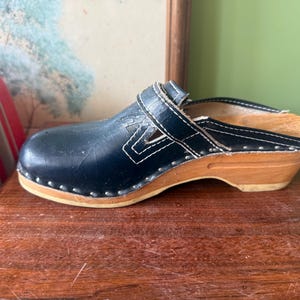 May include: A dark blue leather clog with a wooden sole and a low heel. The upper features a strap across the instep with white contrast stitching and a series of small metal studs securing the leather to the light-colored wooden base.