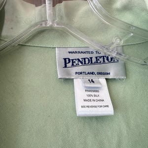 Puede incluir: Primer plano de una camisa de seda verde claro con una etiqueta que dice "Warranted to Pendleton, Portland, Oregon". La etiqueta tambi&eacute;n indica la talla 14 y que la camisa es 100% seda, fabricada en China.