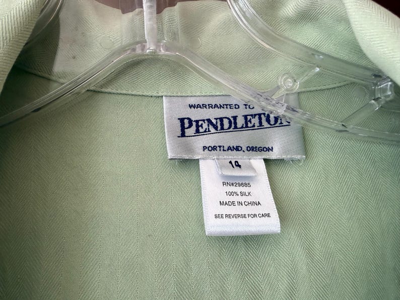 Puede incluir: Primer plano de una camisa de seda verde claro con una etiqueta que dice "Warranted to Pendleton, Portland, Oregon". La etiqueta tambi&eacute;n indica la talla 14 y que la camisa es 100% seda, fabricada en China.